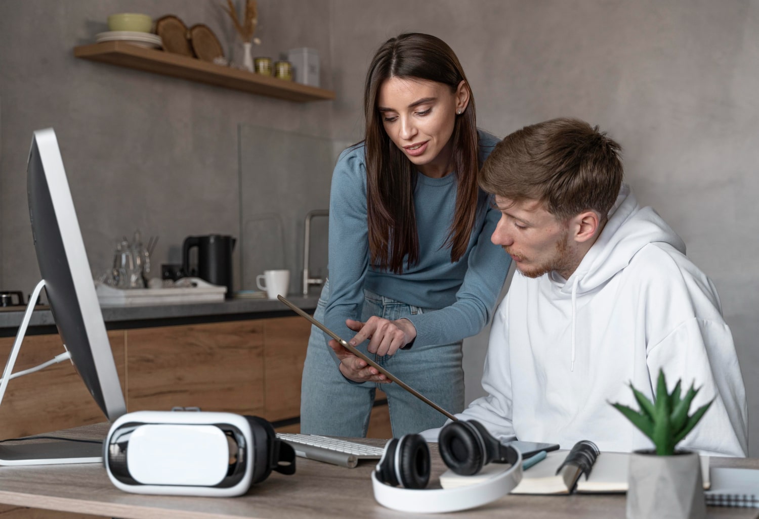A woman and a man review information on a tablet in a modern workspace with a computer, VR headset, and headphones, representing collaboration supported by AI-chatbots.