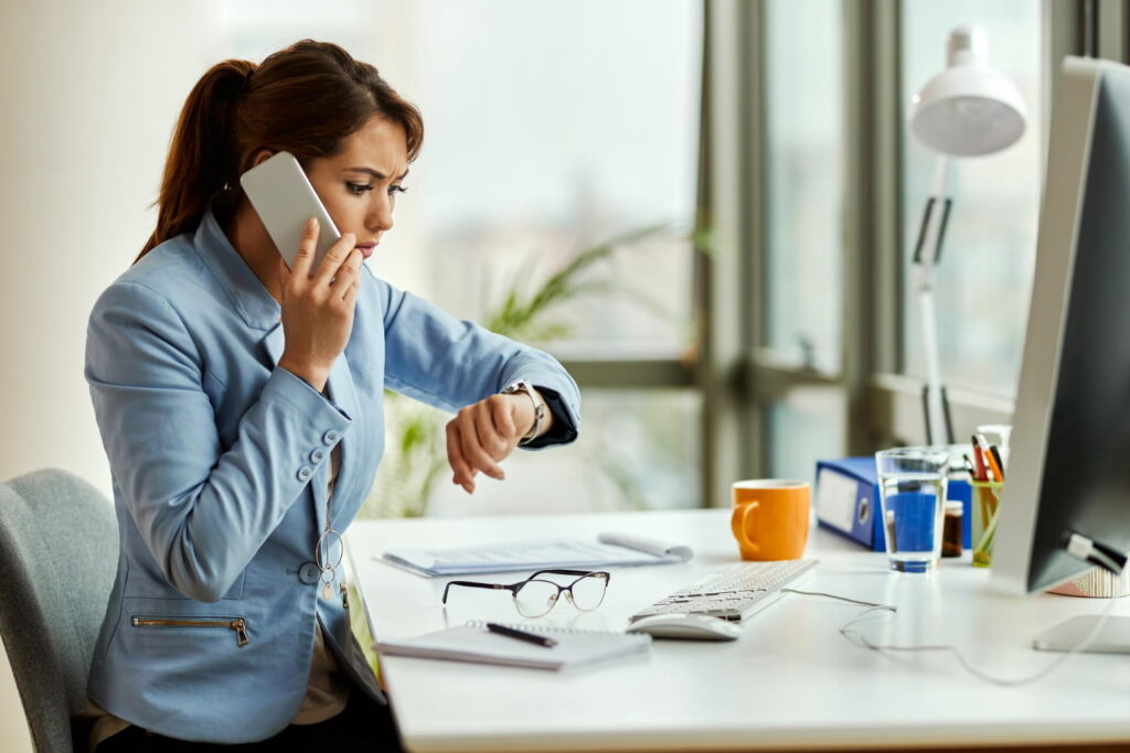 Stressed businesswoman on phone checking watch at desk, highlighting need for appointment booking chatbot solutions